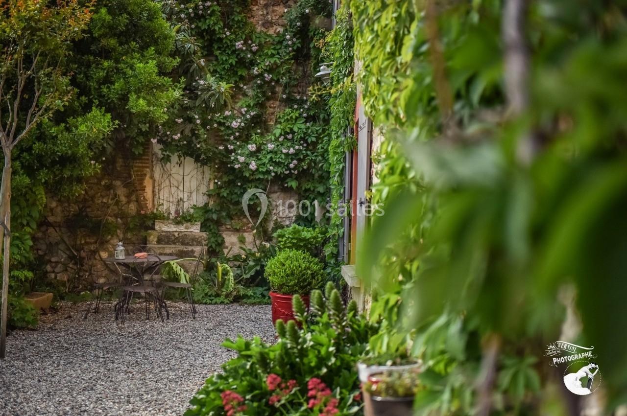 Cour intérieure avec table et chaises en métal, entourée de plantes grimpantes et de pots de fleurs sur un sol en gravier.