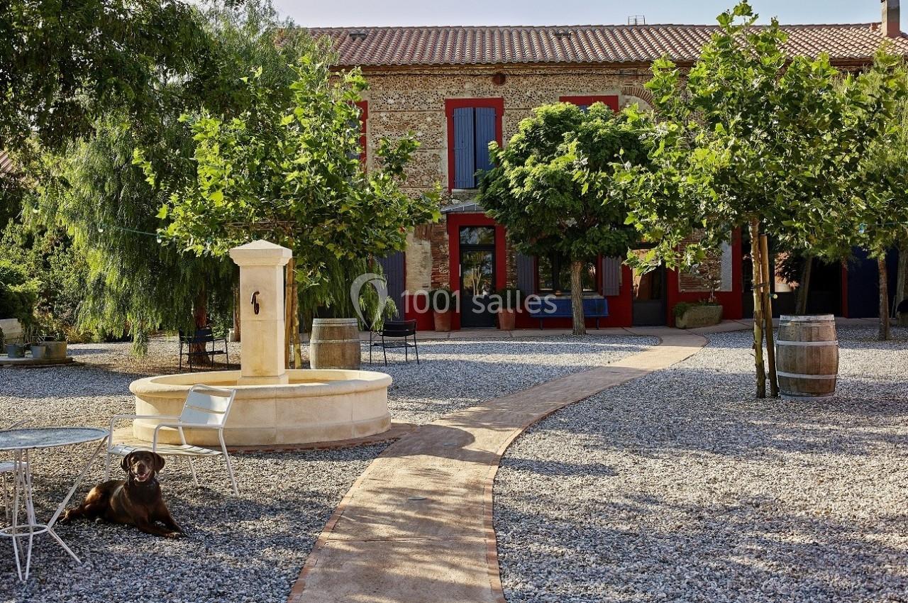 Cour d'une maison en pierre avec une fontaine centrale, des arbres, des tables et un chien allongé sur le gravier.