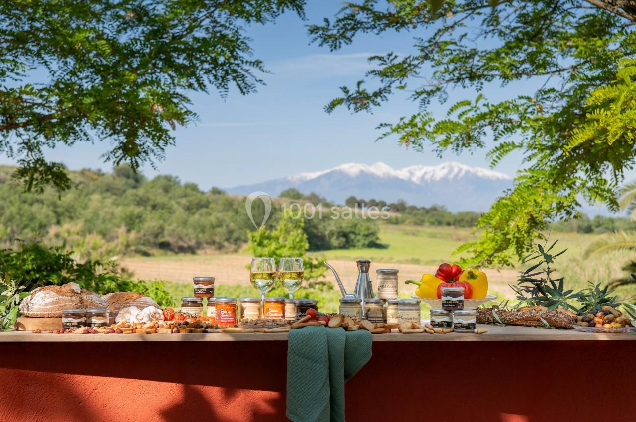 Table garnie de pains, fromages, fruits et condiments, avec vue sur un paysage champêtre et des montagnes en arrière-plan.