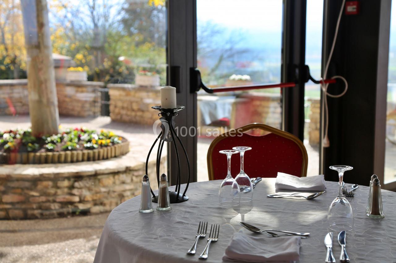 Table dressée avec nappe blanche, verres, couverts et bougeoir, près d'une baie vitrée donnant sur une terrasse ensoleillée.