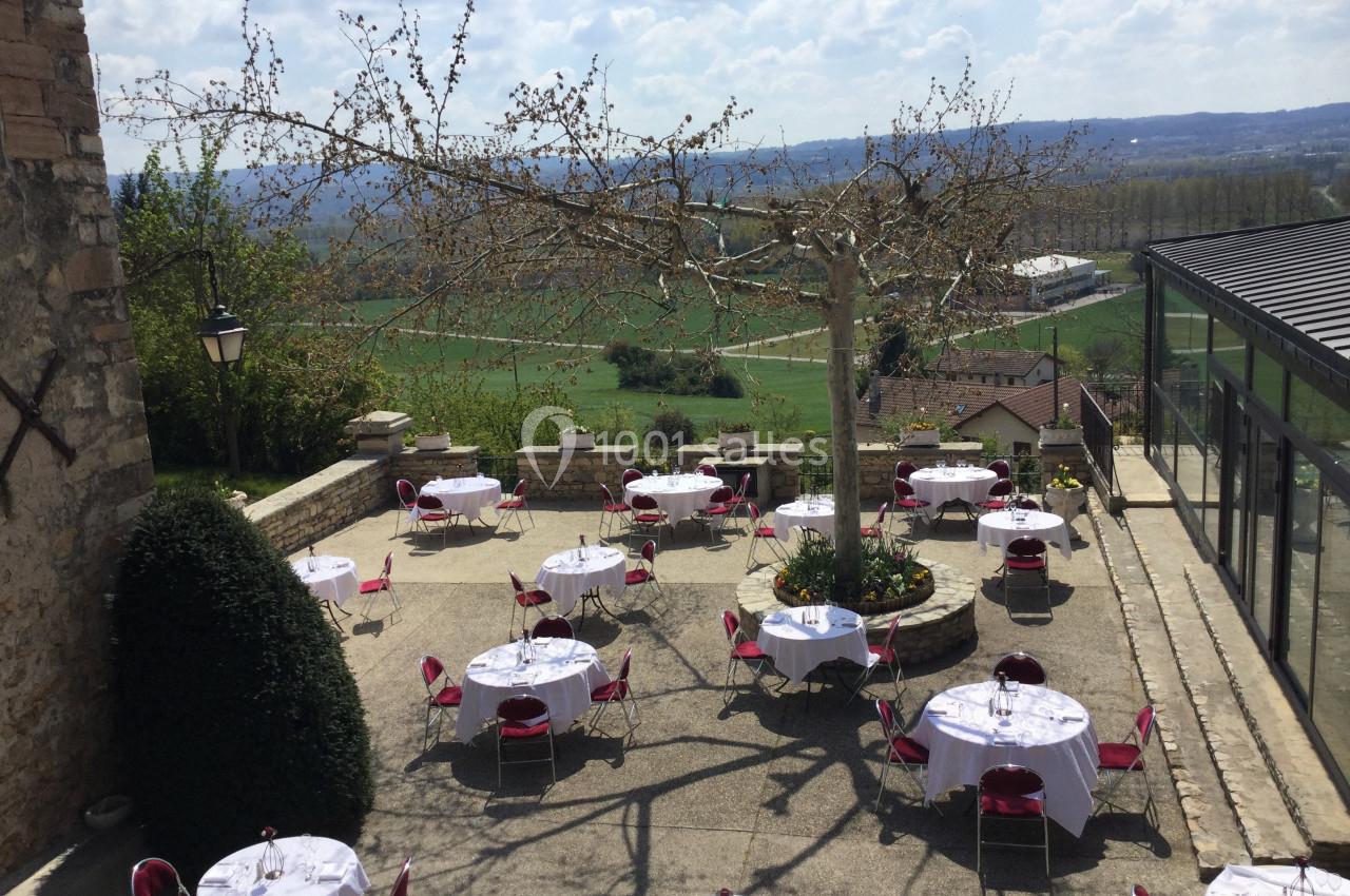 Terrasse extérieure avec tables dressées, chaises rouges, arbre central et vue sur une campagne vallonnée.