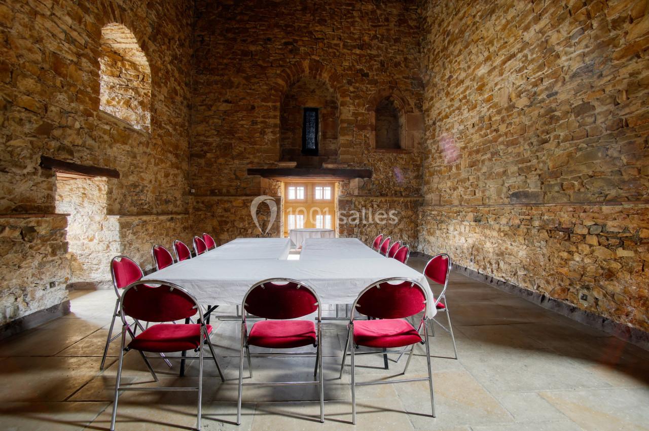 Salle en pierre avec une grande table rectangulaire couverte d'une nappe blanche, entourée de chaises rouges.