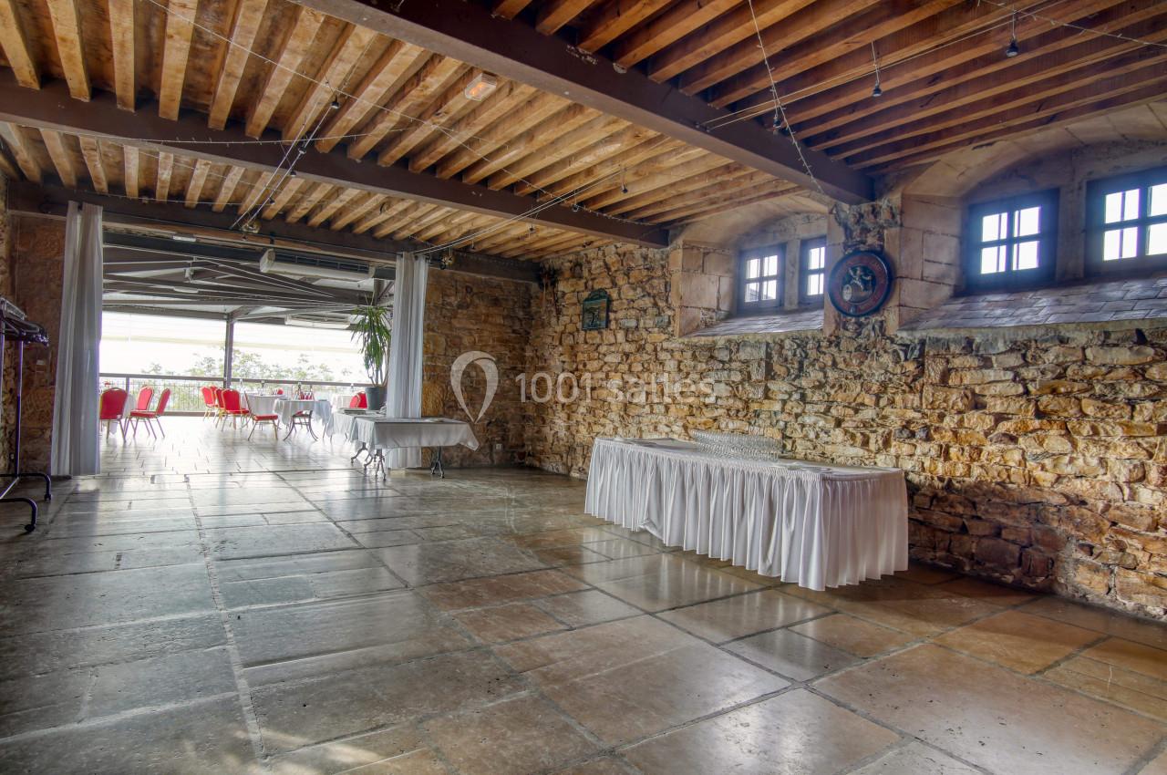 Salle en pierre avec plafond en bois, tables dressées et grandes fenêtres laissant entrer la lumière naturelle.