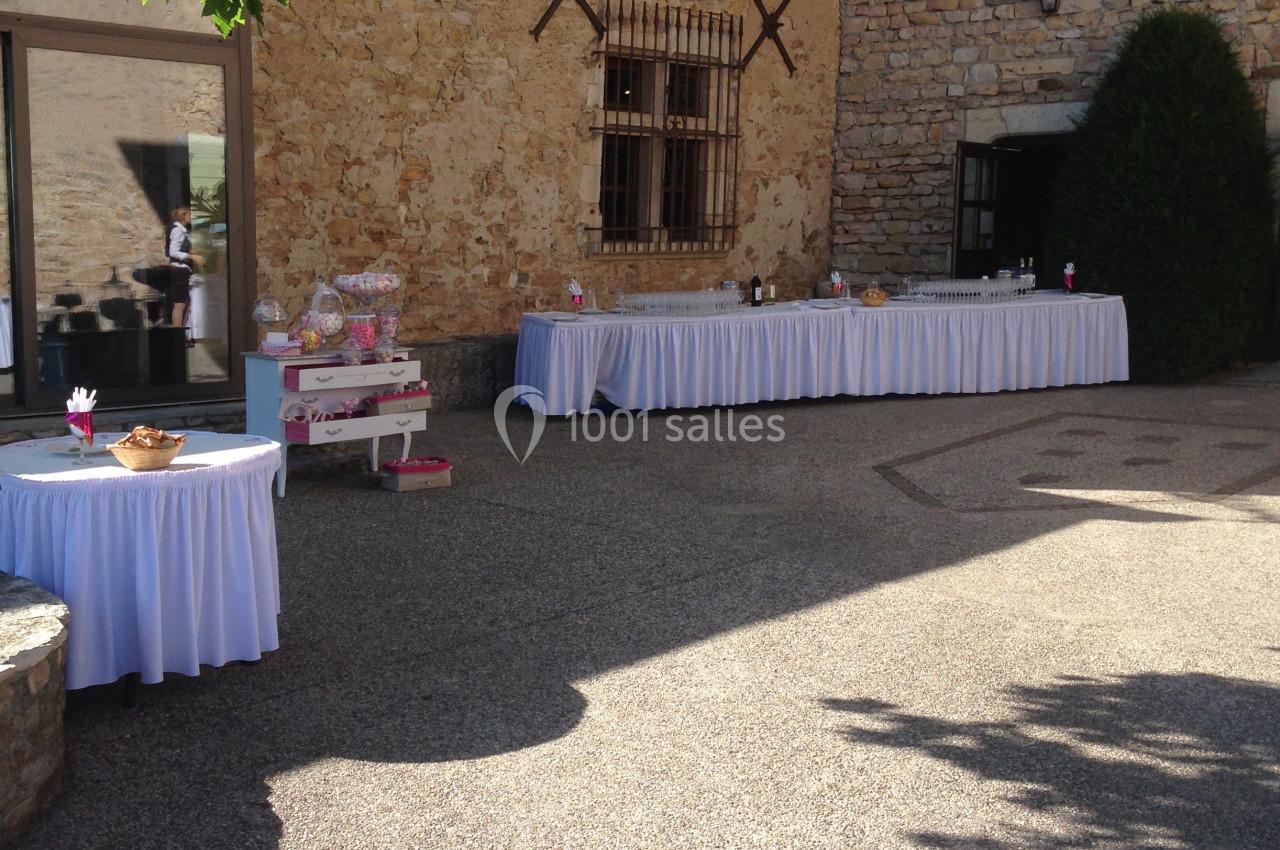 Cour extérieure d'un bâtiment en pierre avec des tables décorées pour un événement, partiellement ombragée par des arbres.