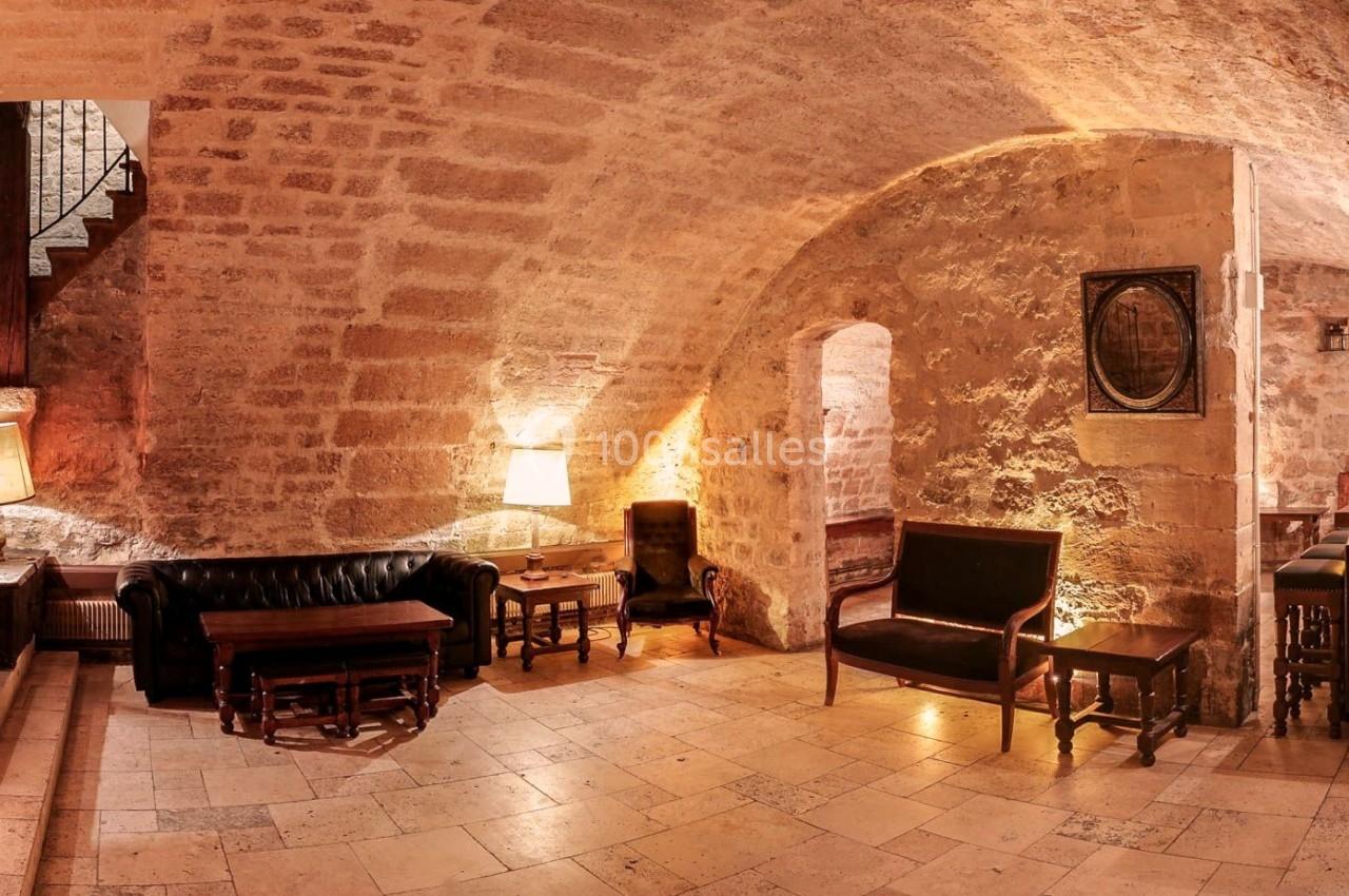 Salon voûté en pierre avec mobilier en cuir, éclairé par des lampes, et un escalier en bois sur la gauche.