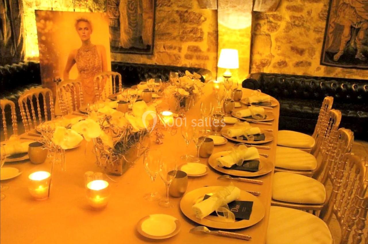 Table élégamment dressée avec nappes dorées, bougies allumées et décorations florales dans une salle en pierre.