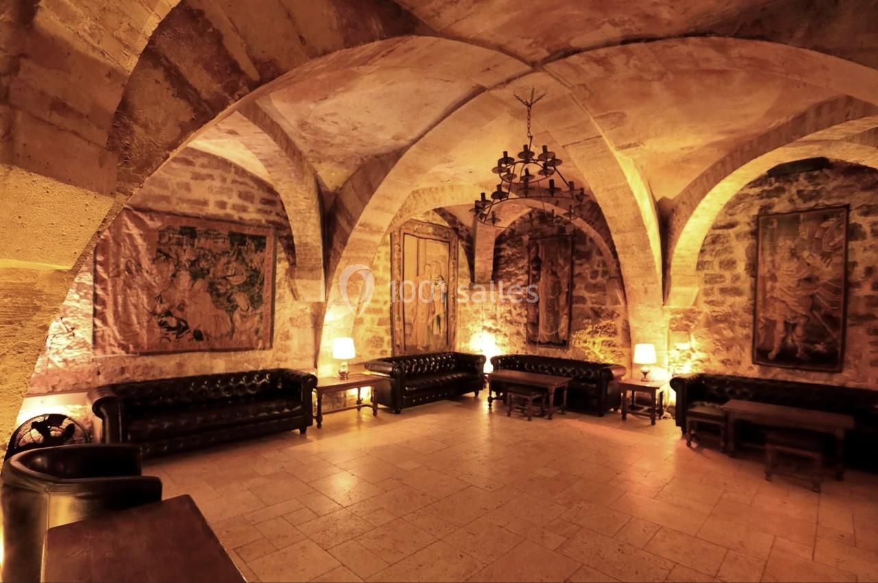 Salle voûtée en pierre avec canapés en cuir, tapisseries murales, éclairage chaleureux et lustre central.