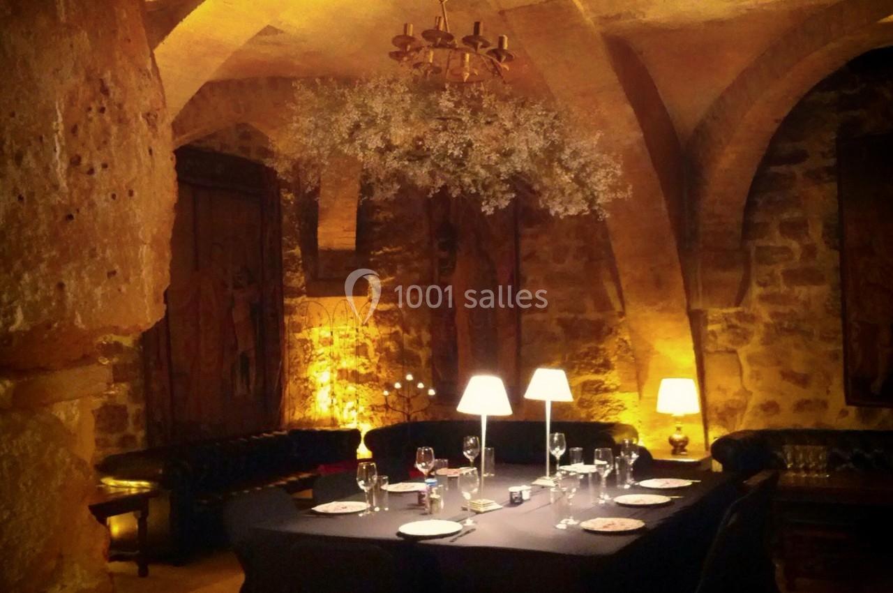 Salle voûtée en pierre éclairée par des lampes et un lustre, avec une table dressée pour un repas.