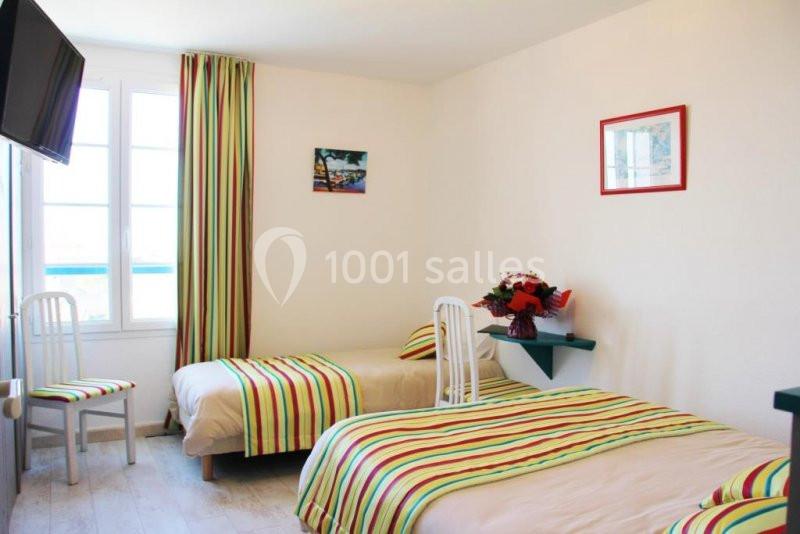 Chambre lumineuse avec deux lits simples, draps rayés colorés, bouquet de fleurs et fenêtre donnant sur l'extérieur.