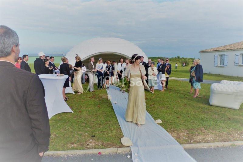 Une femme en robe beige marche sur un tapis blanc lors d'une cérémonie en plein air, entourée d'invités.