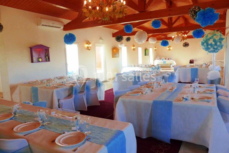 Salle décorée pour un événement avec tables dressées, nappes blanches et chemins de table bleus, ornée de pompons suspendus.