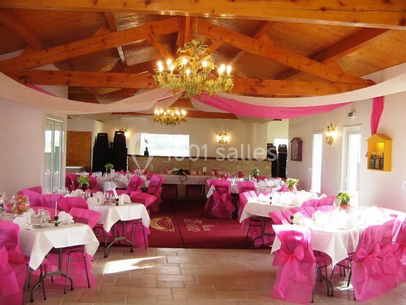 Salle de réception décorée avec des nappes blanches et des nœuds roses sur les chaises, sous un plafond en bois.