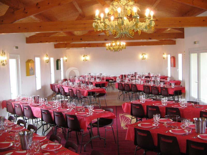 Salle de réception avec tables dressées, nappes rouges, chaises noires et lustres suspendus au plafond en bois.