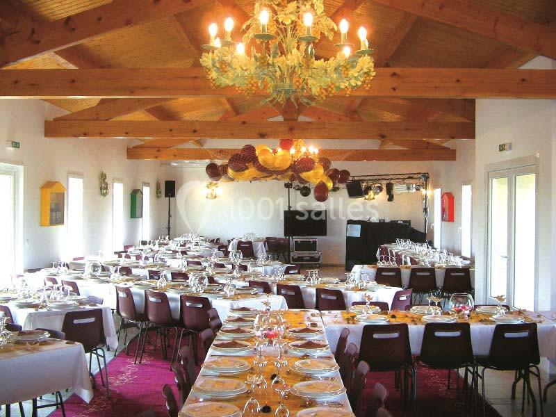 Salle de réception lumineuse avec tables dressées, chaises bordeaux, poutres apparentes et lustre décoratif.