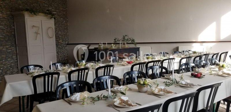 Salle décorée pour un repas festif avec des tables alignées, nappes blanches, vaisselle et arrangements floraux.