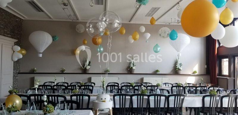 Salle décorée pour une réception avec des tables dressées, des chaises noires et des ballons suspendus aux couleurs vives.