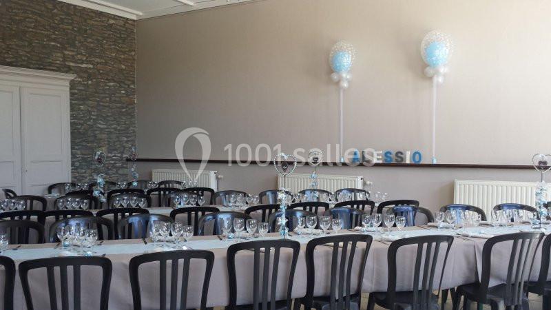 Salle décorée pour un événement avec tables dressées, chaises noires et ballons bleus et blancs en arrière-plan.