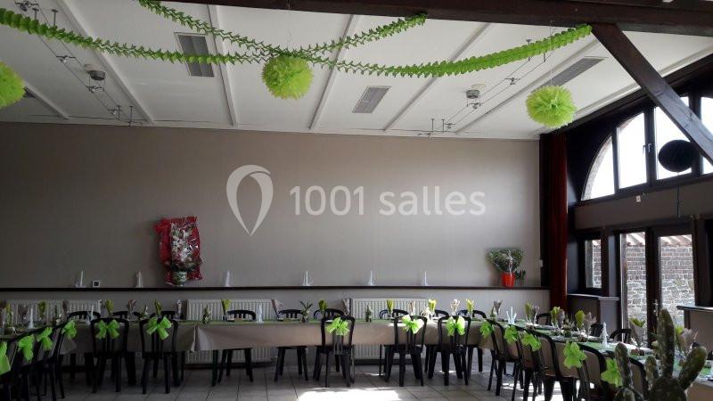 Salle décorée pour un événement, avec des tables alignées, des chaises ornées de nœuds verts et des guirlandes suspendues.