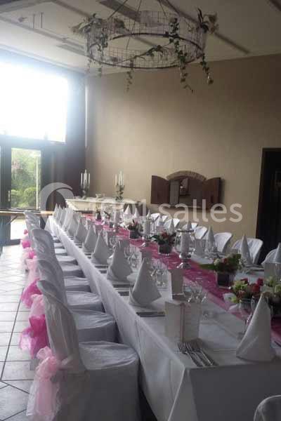 Salle de réception avec une longue table dressée, nappes blanches, serviettes pliées et décorations roses.