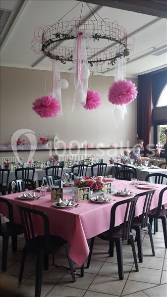 Salle décorée pour une réception, avec tables nappées de rose, chaises noires et suspensions roses et blanches.