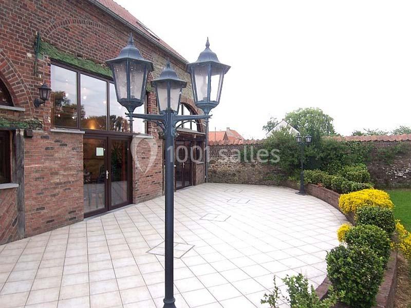 Terrasse pavée avec lampadaires, bordée de buissons et fleurs, adossée à un bâtiment en briques avec grandes fenêtres.
