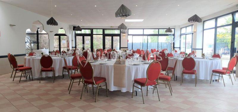 Salle de réception lumineuse avec des tables rondes dressées et des chaises rouges, prête pour un événement.
