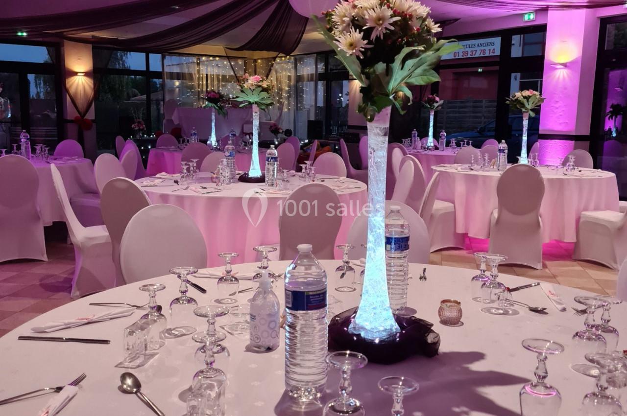 Salle de réception décorée avec des tables rondes, nappes blanches, centres de table fleuris et éclairage rose.