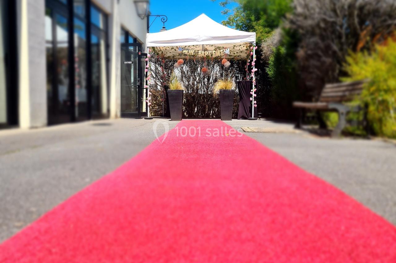 Tapis rouge menant à une tente blanche décorée, entourée de plantes et située dans une allée ensoleillée.