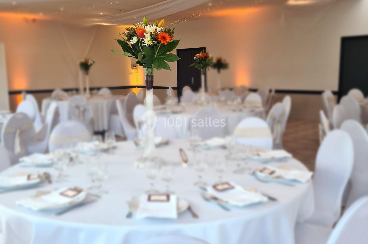 Salle de réception décorée avec des tables rondes drapées de blanc et des centres de table fleuris sous un plafond lumineux.