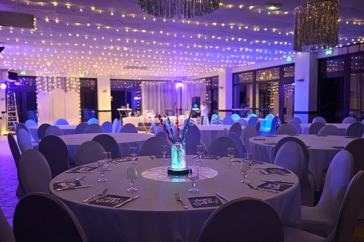 Salle de réception décorée avec des tables rondes, nappes blanches, chandeliers lumineux et guirlandes suspendues.