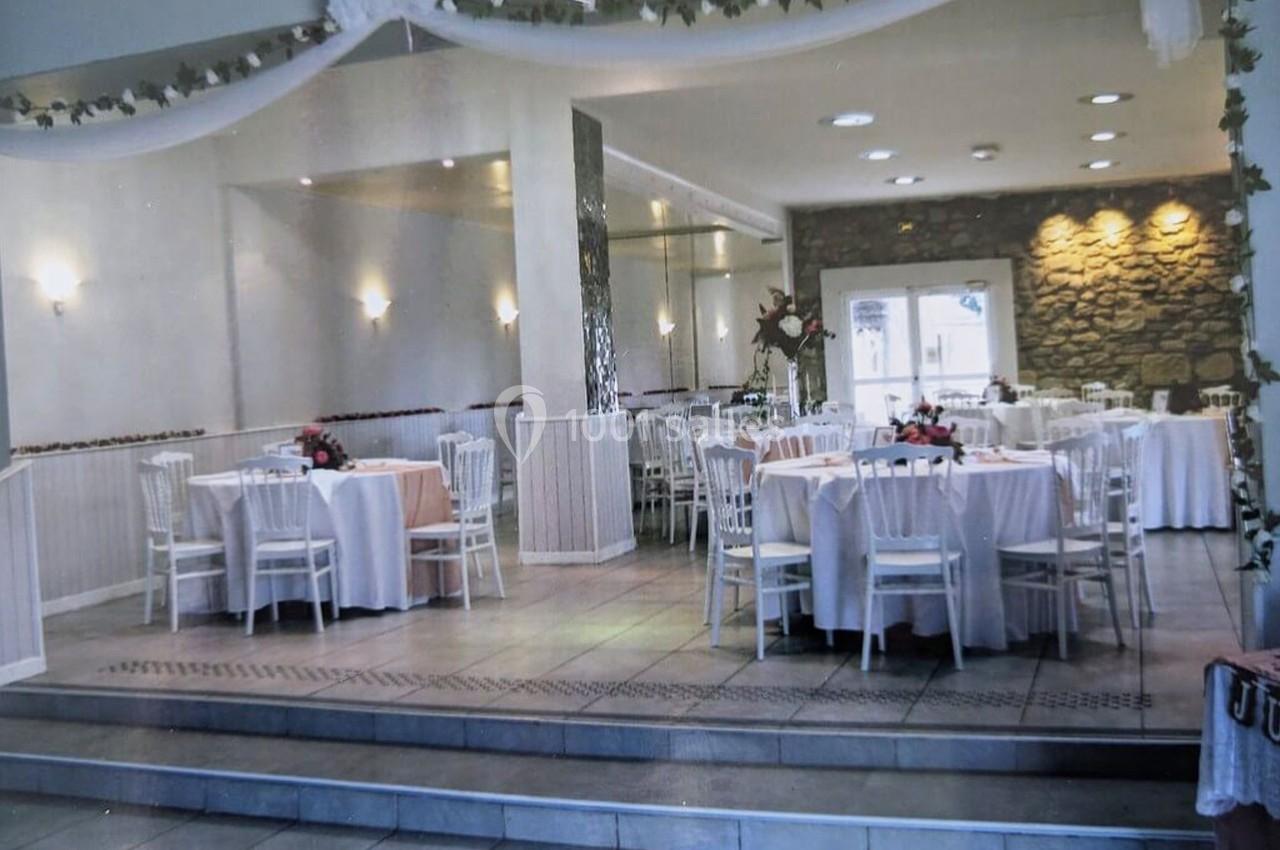 Salle de réception lumineuse avec tables rondes dressées, chaises blanches et décorations florales.