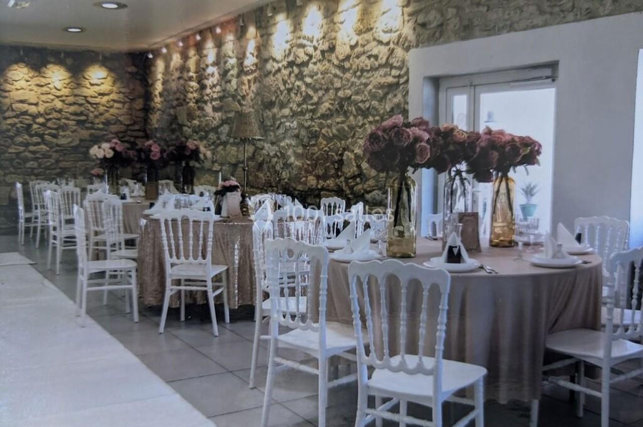 Salle de réception décorée avec des tables rondes, nappes beige, chaises blanches et bouquets de fleurs roses.