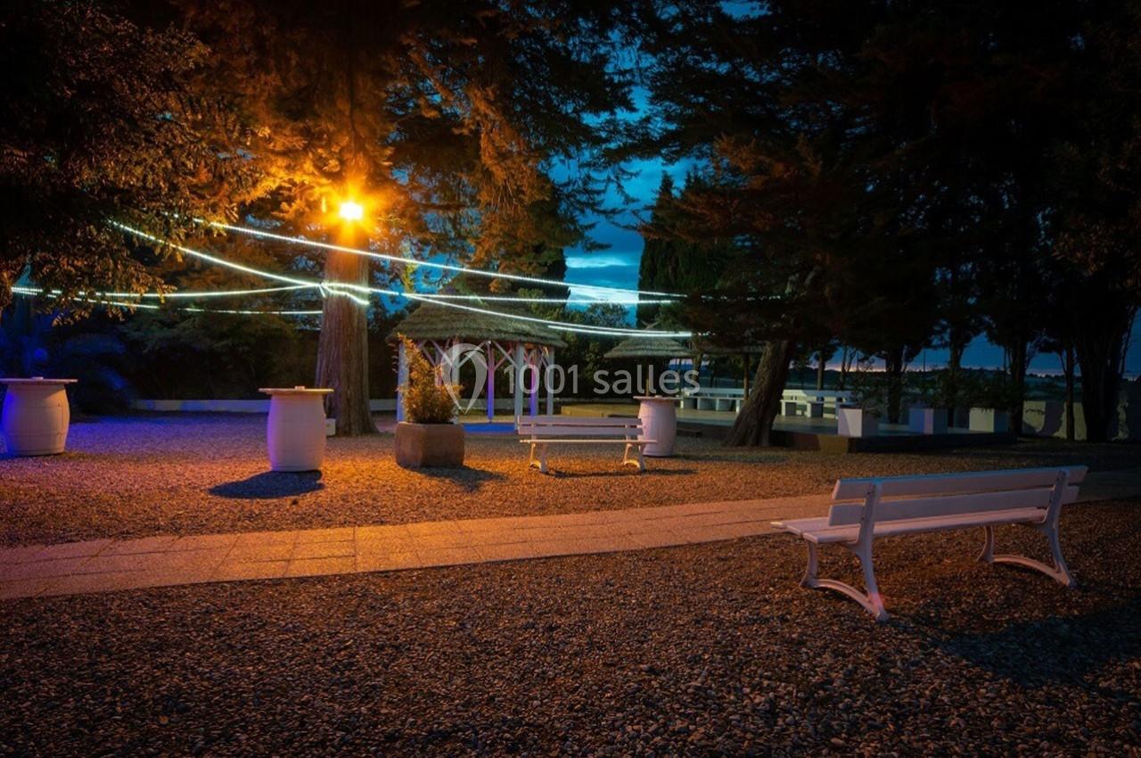 Espace extérieur illuminé avec des guirlandes lumineuses, bancs blancs et arbres entourant une pergola.