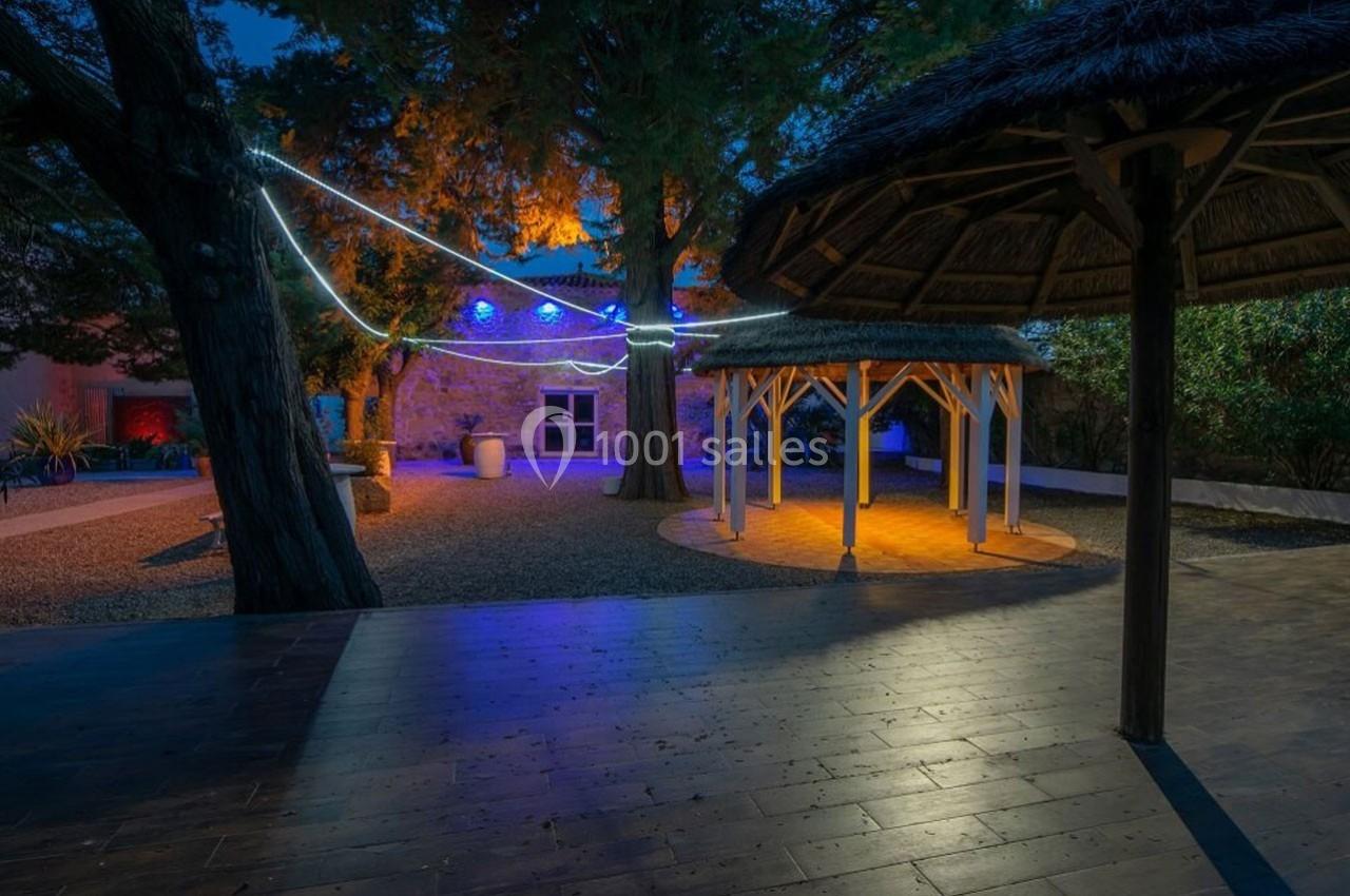 Cour extérieure éclairée de nuit avec des pergolas en bois, des guirlandes lumineuses et des arbres environnants.