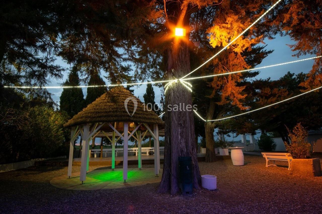 Abri en bois éclairé par des lumières colorées, entouré d'arbres et de guirlandes lumineuses dans un espace extérieur.