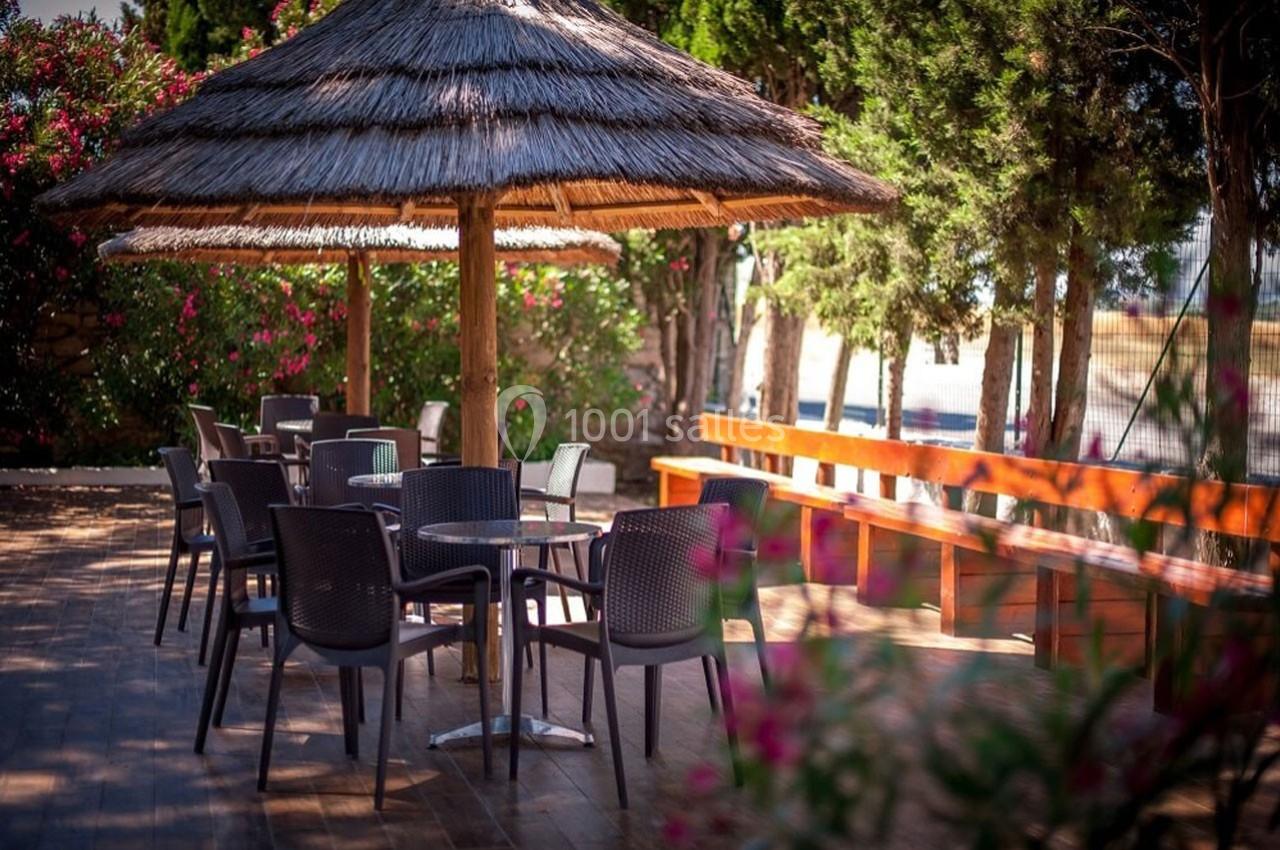Terrasse en bois avec tables et chaises sous un parasol en chaume, entourée de végétation et de fleurs colorées.