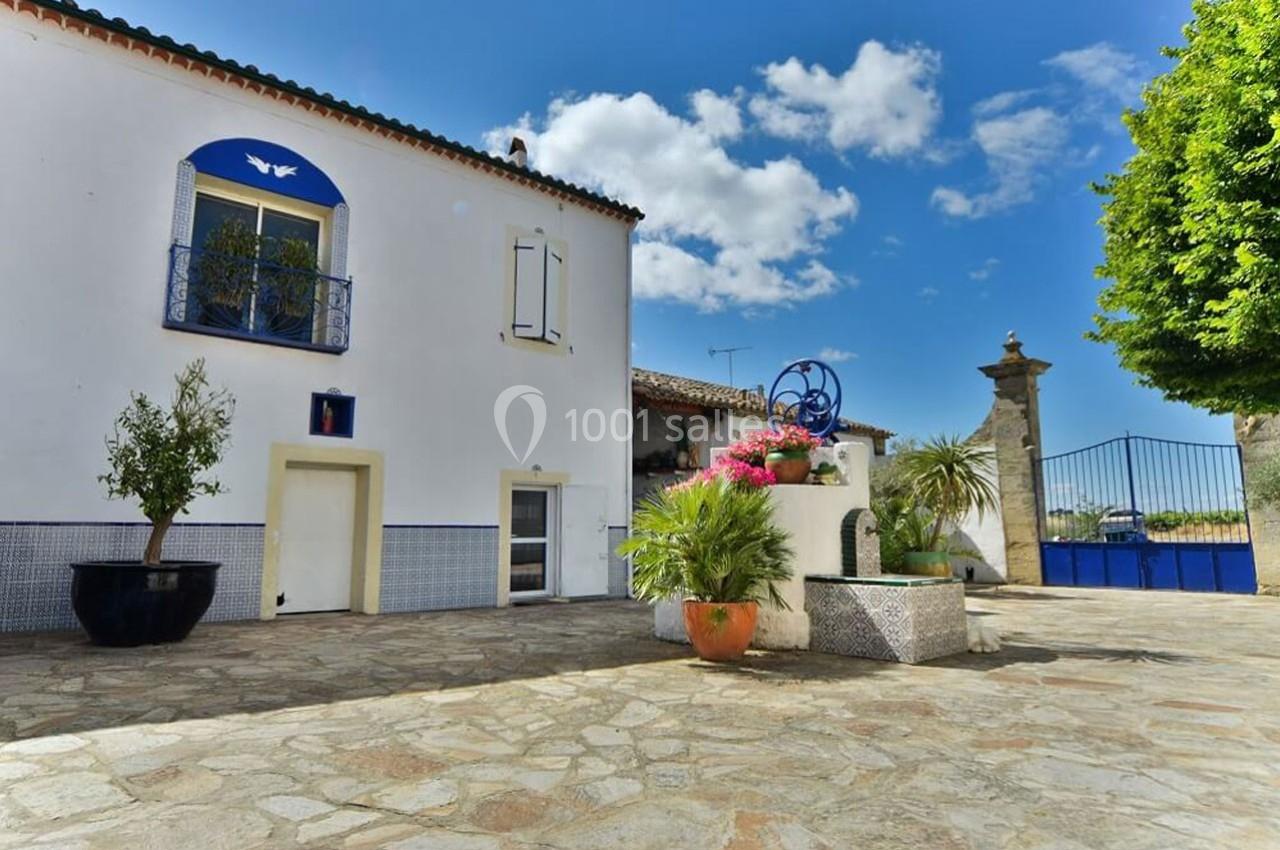 Cour pavée avec une maison blanche, des plantes en pot, une fontaine décorative et un ciel bleu en arrière-plan.