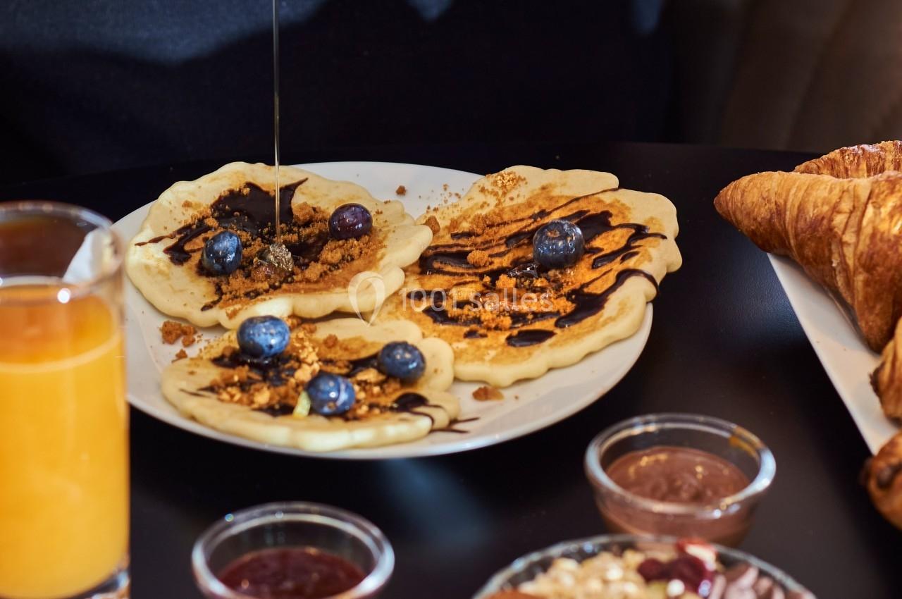 Une main verse du miel sur des pancakes garnis de myrtilles et de noix, entourés de divers aliments pour le petit-déjeuner.