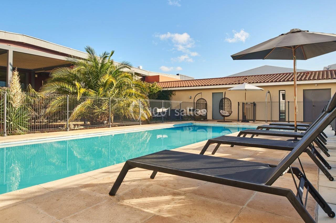 Piscine extérieure entourée de transats noirs, parasols et végétation, devant un bâtiment moderne sous un ciel bleu.