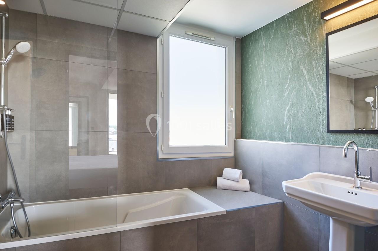 Salle de bain moderne avec baignoire, lavabo, miroir, fenêtre et murs en carrelage gris et vert.