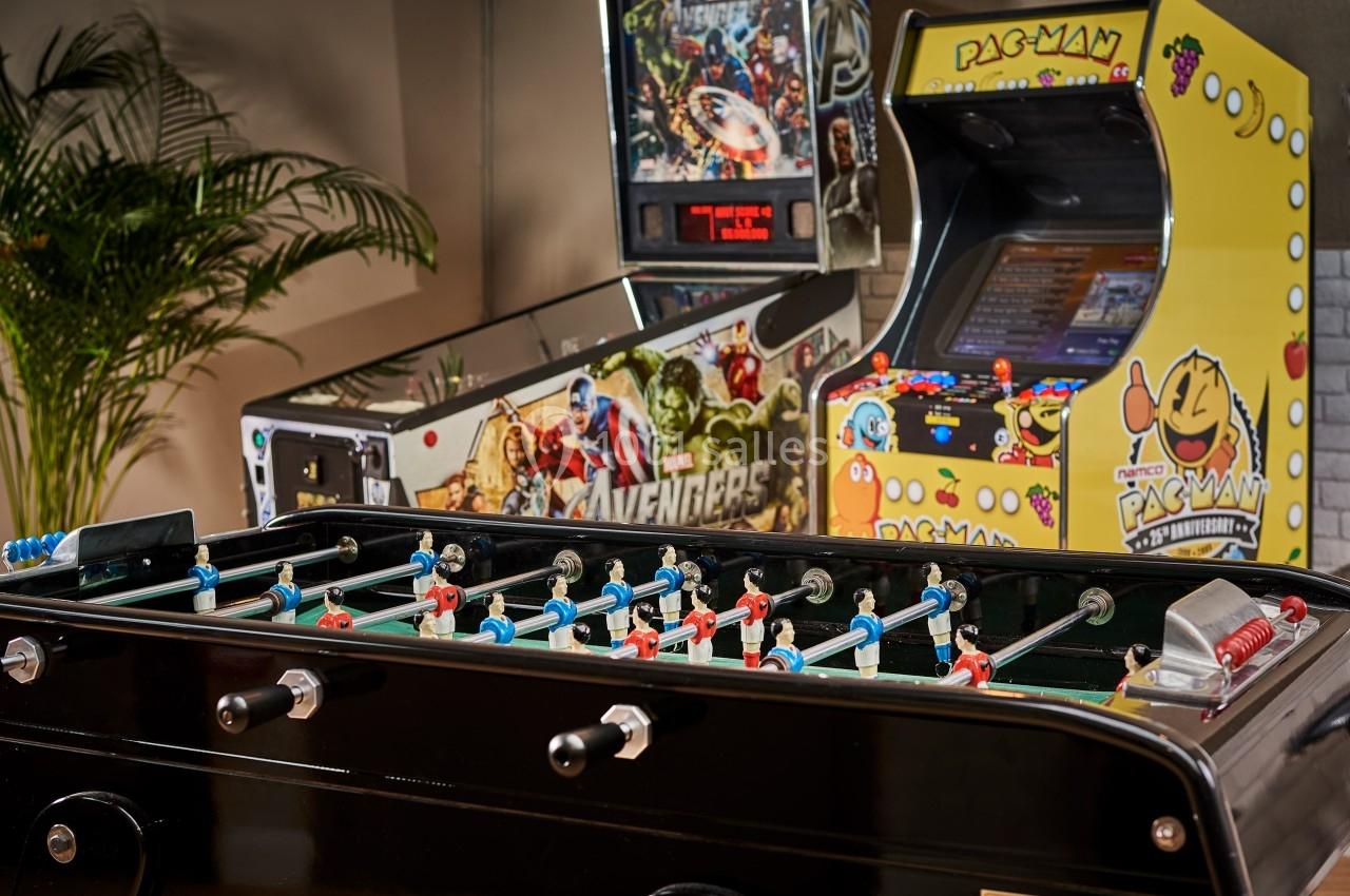 Baby-foot noir au premier plan, avec en arrière-plan des bornes d'arcade Avengers et Pac-Man dans une pièce lumineuse.