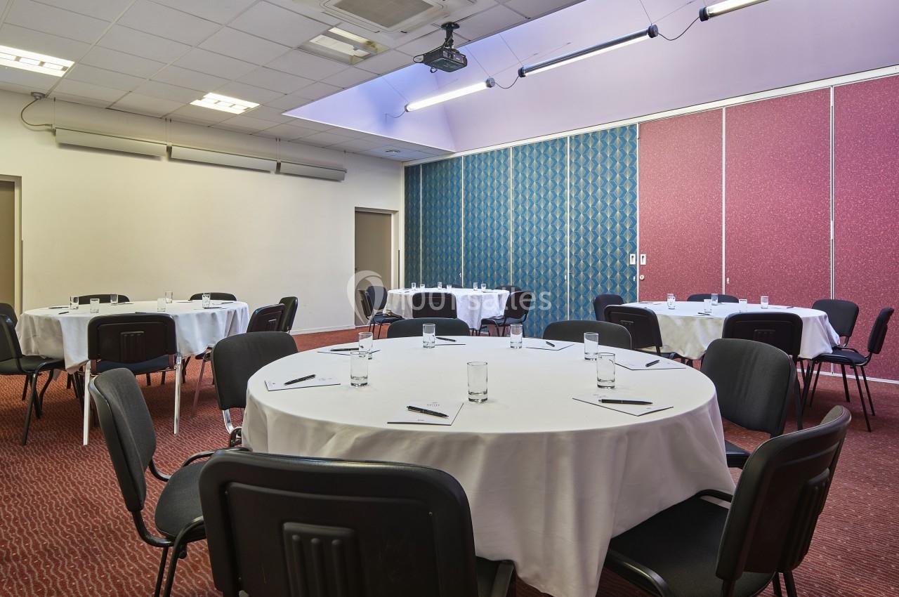 Salle de réunion avec tables rondes dressées de nappes blanches, chaises noires et fournitures de bureau.