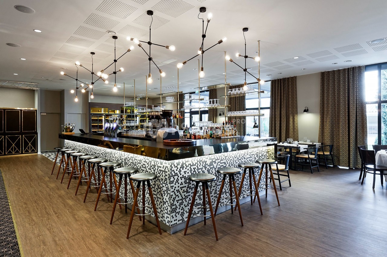Bar moderne avec comptoir en mosaïque, tabourets alignés, éclairage suspendu et tables en arrière-plan.