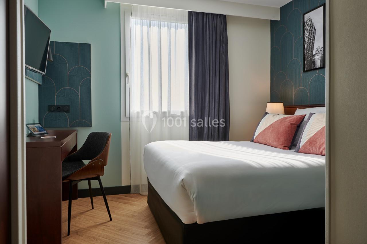 Chambre d'hôtel lumineuse avec un lit double, un bureau, une chaise, une télévision murale et une fenêtre avec rideaux.