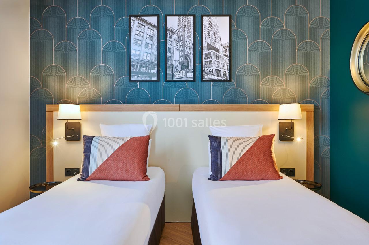 Deux lits simples avec coussins colorés, tête de lit en bois clair, lampes murales et décor mural bleu avec cadres.