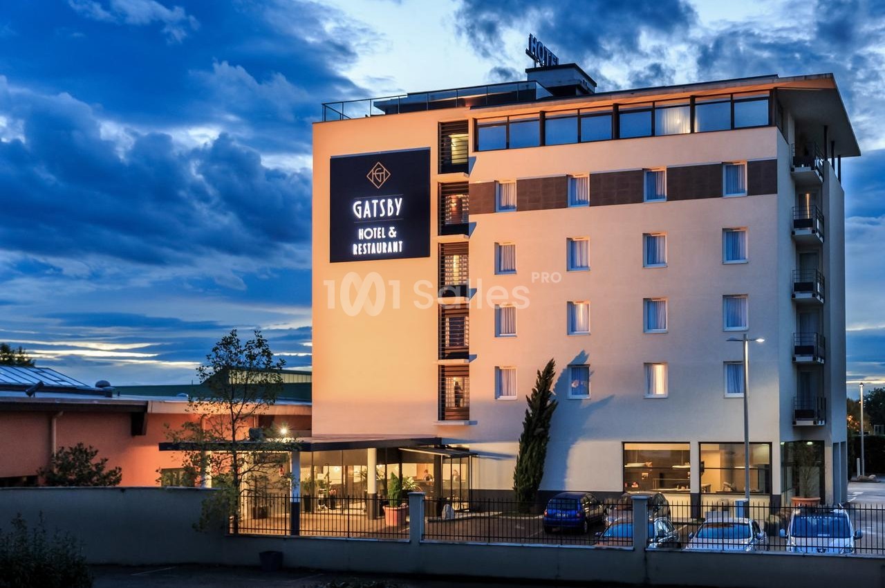 Façade d'un hôtel moderne de plusieurs étages avec enseigne lumineuse, entouré de quelques arbres et voitures stationnées.