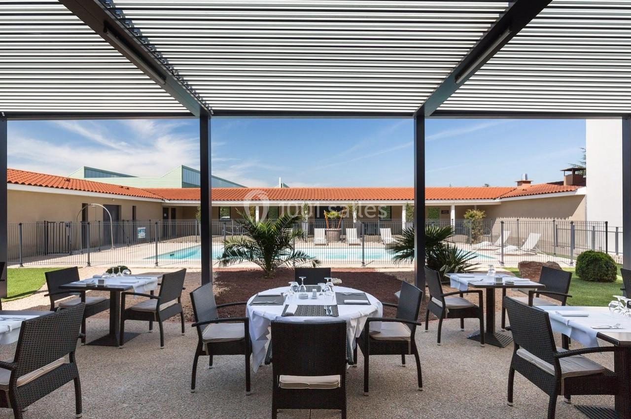 Terrasse aménagée avec des tables et des chaises, donnant sur une piscine entourée de végétation et de bâtiments.