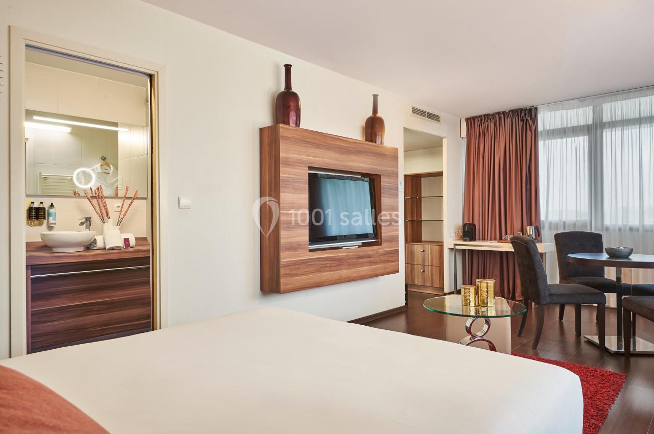 Chambre moderne avec lit double, télévision murale, coin repas, et salle de bain visible à travers une porte ouverte.