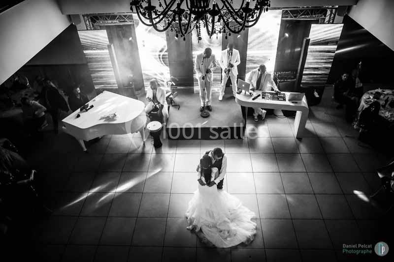 Un couple danse sous un lustre dans une salle de réception, avec un groupe de musiciens en arrière-plan.