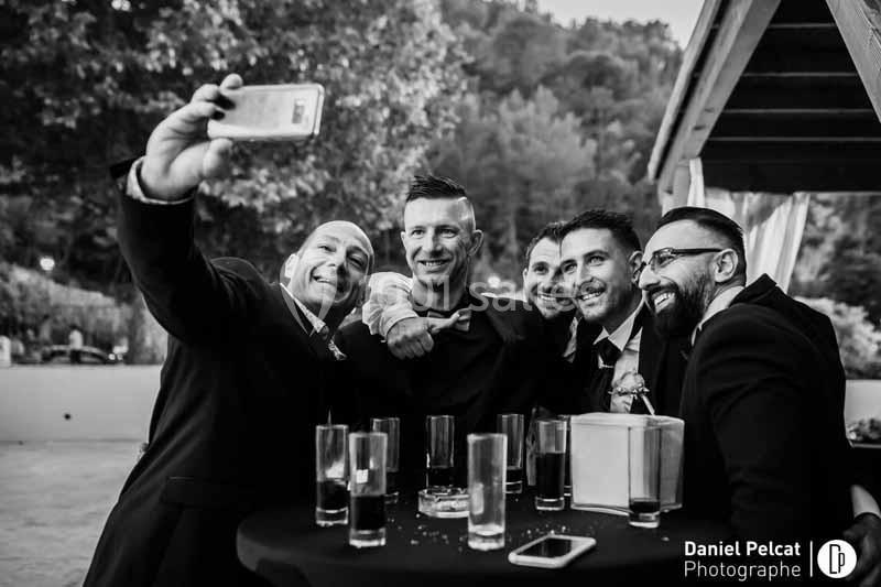 Des hommes en costume sourient et prennent un selfie autour d'une table avec des verres, en extérieur.
